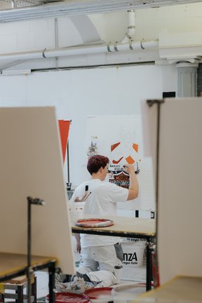 Eine Person kniet beim Bemalen einer Leinwand in einem Kunstatelier, umgeben von anderen Leinwänden und Kunstzubehör.