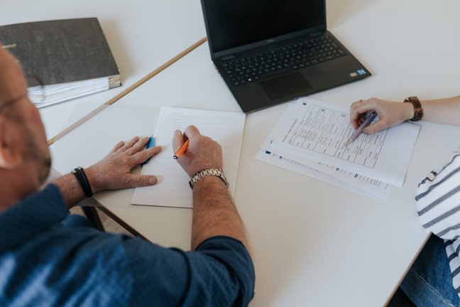 Zwei Personen arbeiten an einem Schreibtisch mit einem Laptop, einem Notebook und Dokumenten, schreiben und prüfen Papiere.