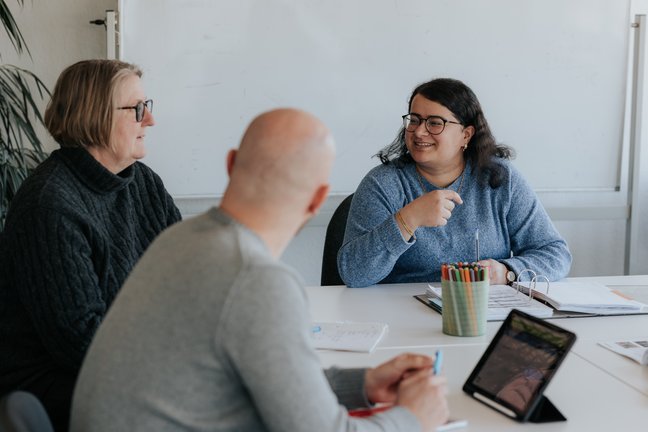 Drei Personen sitzen in einem Besprechungsraum um einen Tisch herum und unterhalten sich, wobei sie Papiere und ein Tablet vor sich haben.