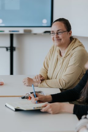 Eine Frau im hellbraunen Kapuzenpulli sitzt lächelnd an einem Tisch mit Notizbüchern und Stiften während einer Besprechung.
