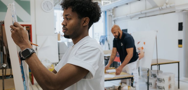 Ein Mann malt auf einer Leinwand in einem hellen Atelier, während ein anderer Mann im Hintergrund arbeitet.