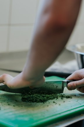 Person, die in einer Küche frische Kräuter auf einem grünen Schneidebrett mit einem großen Messer hackt.