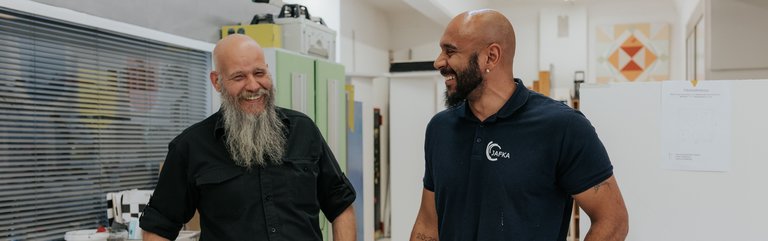 Zwei Männer in Arbeitskleidung stehen drinnen, lächeln und unterhalten sich in einem Raum, der eine Werkstatt oder ein Atelier zu sein scheint.