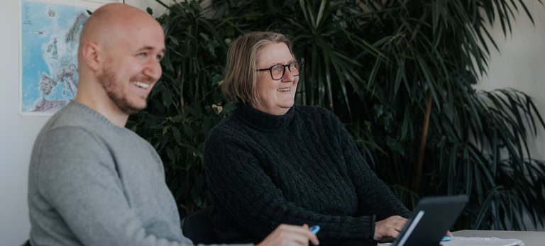 Zwei lächelnde Menschen sitzen an einem Tisch mit Papieren und einem Tablet, vor großen Grünpflanzen.