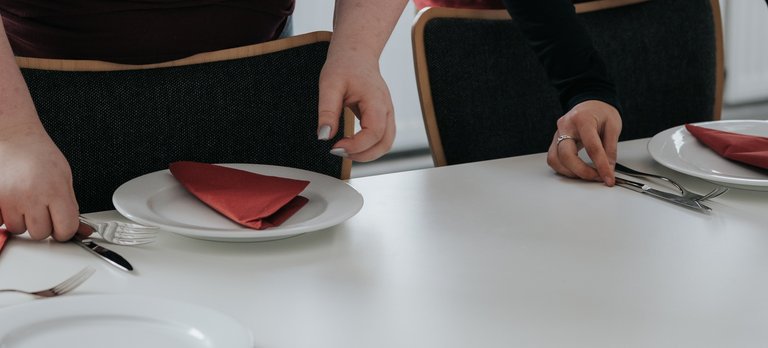 Zwei Frauen in weinroten Hemden decken einen Tisch mit Tellern, Servietten und Besteck.