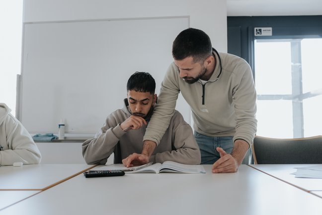 Ein Lehrer hilft einem Schüler bei seiner Arbeit an einem Schreibtisch in einem Klassenzimmer und zeigt auf ein aufgeschlagenes Buch.