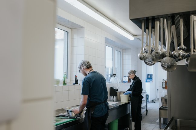 Zwei Personen mit Haarnetzen und Handschuhen arbeiten an den Spülbecken in einer sauberen, hellen Großküche.