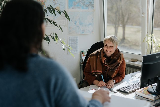 Eine Frau lächelt eine Person an, die in einem hellen Büro mit Karten an der Wand sitzt.