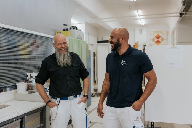 Zwei Männer stehen und lächeln sich in einer hellen, modernen Werkstatt- oder Studioumgebung an.