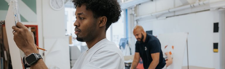 Ein Mann malt auf einer Leinwand, während ein anderer im Hintergrund in einem hellen Kunstatelier arbeitet.