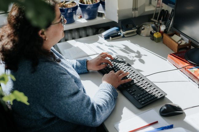 Eine Person tippt auf einer Tastatur an einem Schreibtisch mit Büromaterial und Pflanzen im Hintergrund.