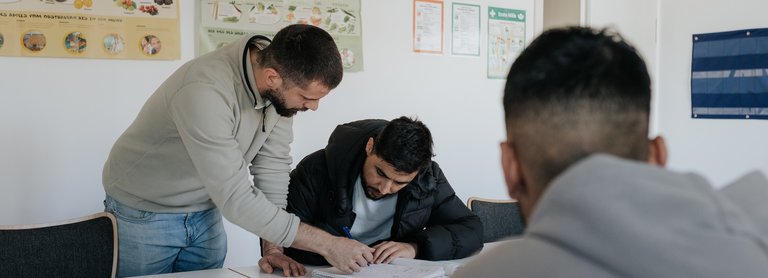 Ein Mann hilft einem anderen Mann an einem Tisch mit Papierkram, während eine dritte Person im Vordergrund sitzt.