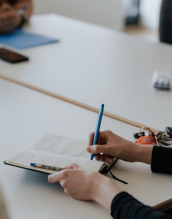 Person, die während einer Besprechung an einem Tisch in ein Notizbuch schreibt, mit Schlüsseln und einem Stift in der Nähe.
