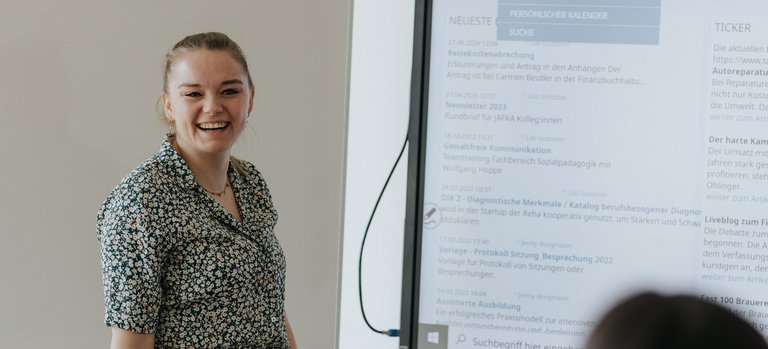 Eine Frau lächelt, während sie vor einem Bildschirm und einem Whiteboard vor einem sitzenden Publikum präsentiert.