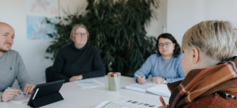 Vier Personen sitzen in einer Besprechung um einen Tisch herum, besprechen Dokumente und machen Notizen.